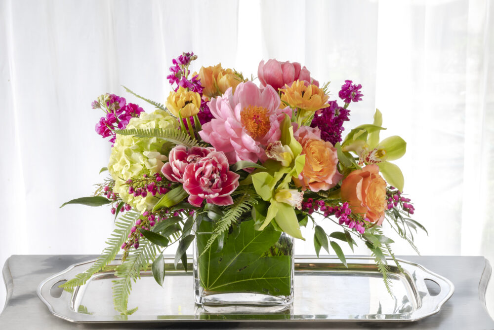 Vibrant coral peonies and roses with bright magenta spring flowers arranged in an emerald leaf wrap for a unique Mother’s Day gift, delivered.
