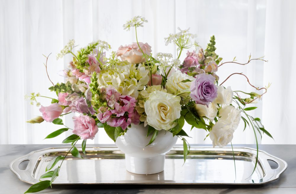 A lovely feminine design of soft pink, lavender, and white blooms in a striking pedestal bowl, perfect for a gift, delivered.