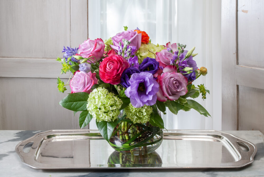 Lively and bright flowers in bold pink, purple, and fresh spring green designed in an emerald wrapped rose bowl for a lovely gift, delivered.
