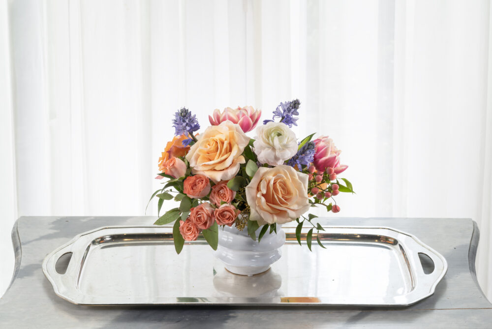 Cheerful fresh flower arrangement in peach, pink, and lavender blooms designed in a white ceramic pot for a Mother’s Day gift.