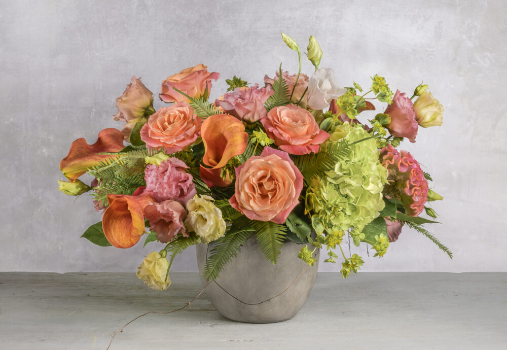 A bright coral and pink fresh flower arrangement with colorful orange callas in a smooth grey pot.