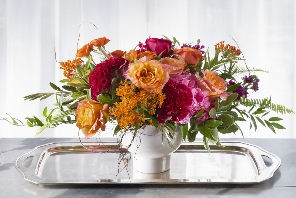 Vibrant summer fresh arrangement with beautiful roses and field flowers in bold pinks and orange, designed in a chic white pedestal bowl.