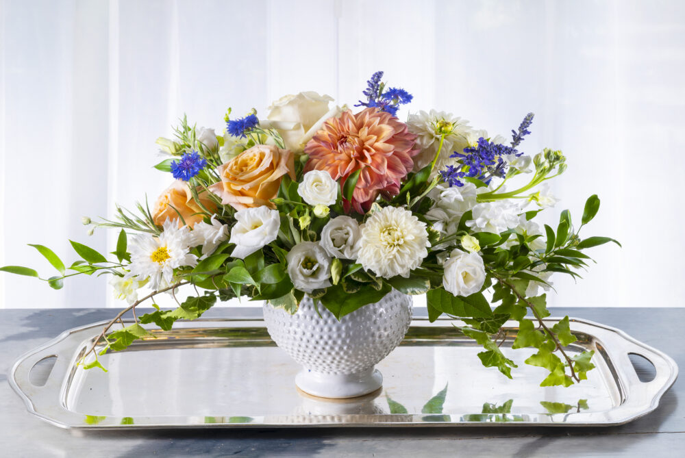 A lovely, light fresh flower arrangement of summer peach, coral, and white with blue accents, Designed in a white cachepot, delivered.