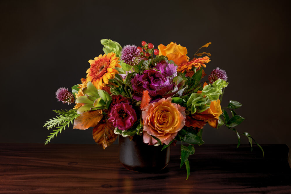Vibrant autumn colors in orange, coral, purple, and amber with accents of autumn leaves. Fresh seasonal flowers arranged a matte black pot for a bright display.