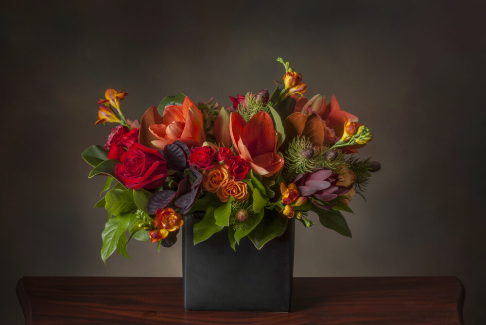 Unique taller vibrant seasonal fresh flower arrangement with rich colors of orange, red, and wine in a slim matte black rectangle.