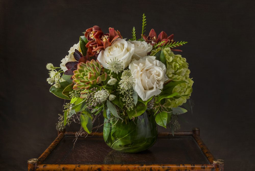 Unique autumn arrangement with deep red orchids and succulents with crisp white roses in an emerald leaf wrap, delivered.