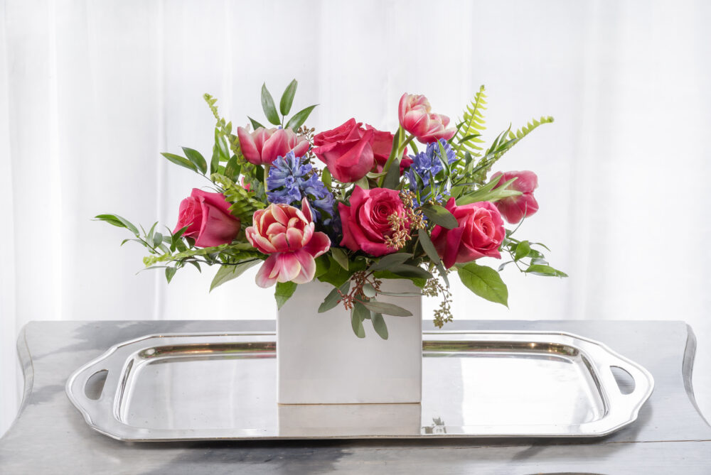 Bold and bright pink roses and spring tulips with blue hyacinth flowers arranged in a slim white matte rectangle.