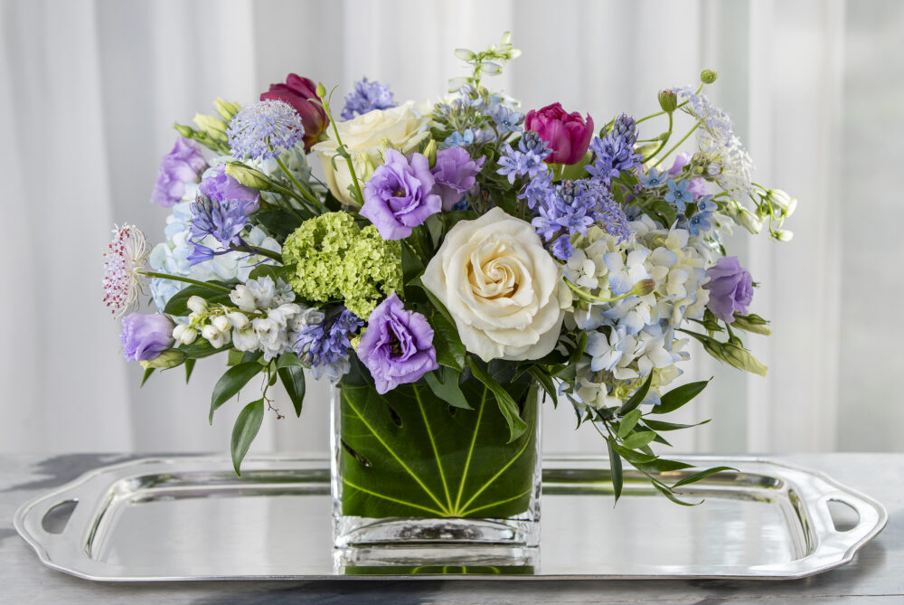 A lush Mother’s Day gift, fresh spring flower bouquet of bright blue, lavender, and white with spring green foliage in a leaf-wrapped rectangle, delivered.