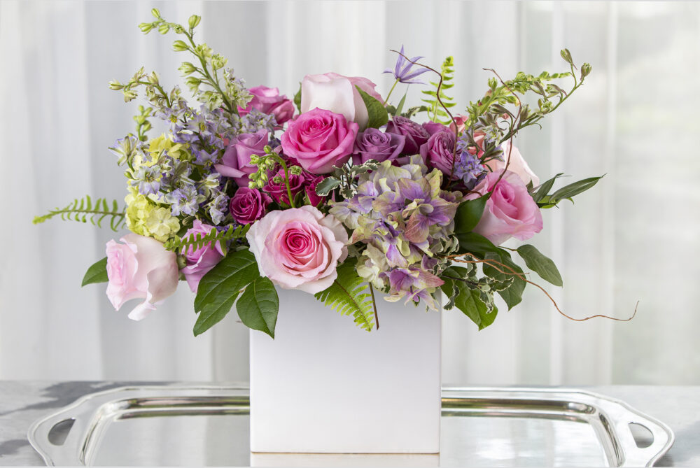 Lush and lovely roses in pink, purple, and lavender with spring flowers in a higher design. delivered for you.
