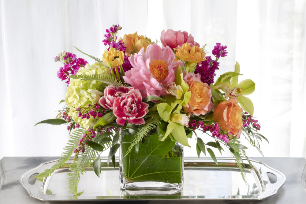 Vibrant coral peonies and roses with bright magenta spring flowers arranged in an emerald leaf wrap for a unique Mother’s Day gift, delivered.