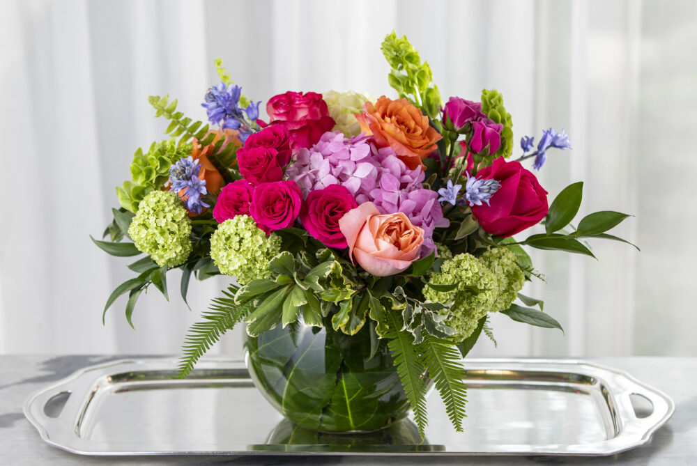 Lively and bright flowers in bold pink, purple, coral, and fresh spring green designed in an emerald wrapped rose bowl for a lovely gift, delivered.