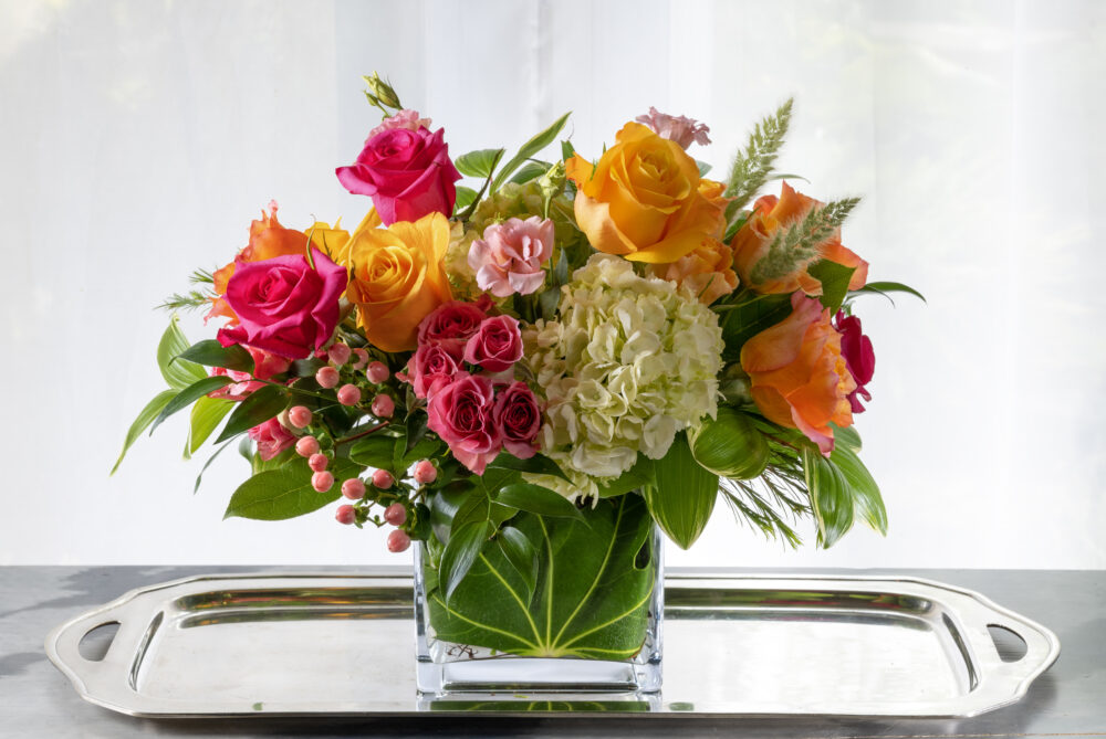 A vibrant arrangement in fresh flowers of coral, bold pink, and orange in a colorful display. Designed in an emerald leaf-wrapped rectangle, delivered.