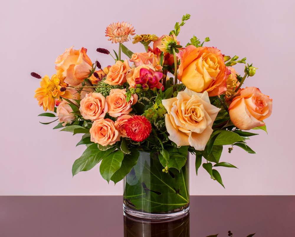 A bright peach mix of autumn fresh flowers with peach, orange, and pink blossoms in a glass cylinder vase, delivered.