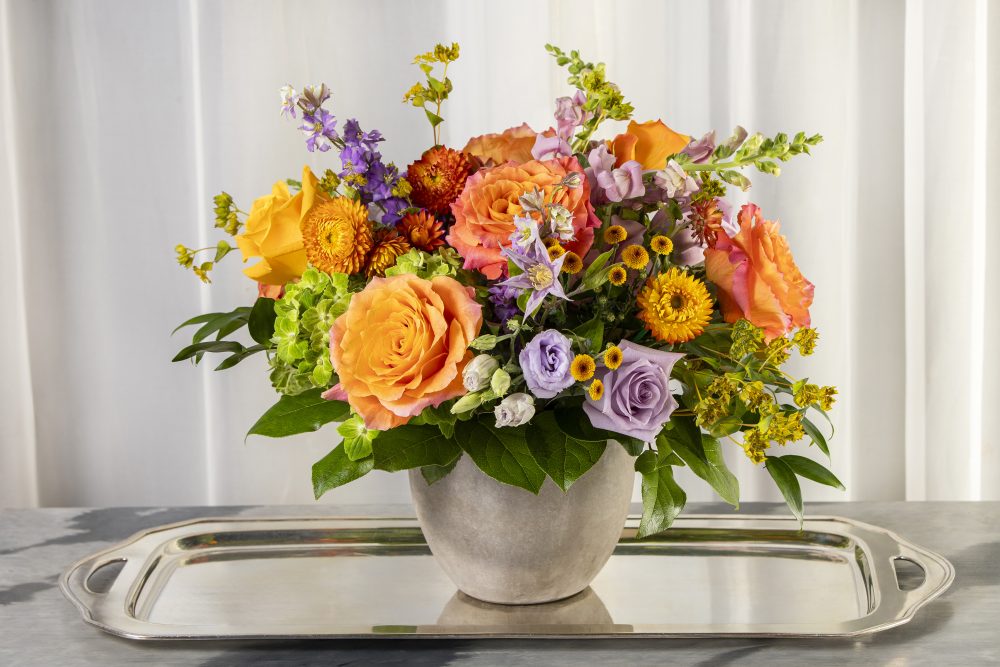 A vibrant mix of summer fresh flowers with coral, orange, and lavender, with yellow accents in a smooth grey pot for a cheerful gift, delivered.