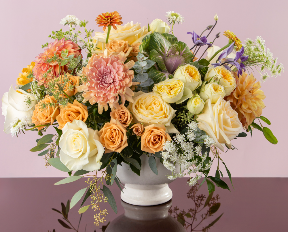 A pastel mix of autumn fresh flowers with peach, green, yellow, white and pink blossoms in a ceramic compote vase, delivered.