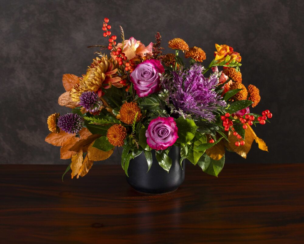 A vibrant purple and orange mix of autumn fresh flowers with purple and orange blossoms and seasonal foliage in a black ceramic cylinder, delivered, thanksgiving.
