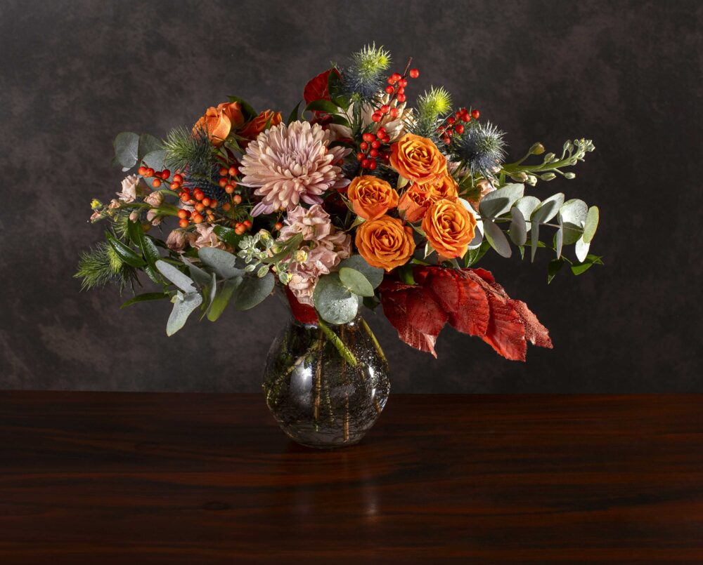 A seasonal mix of autumn fresh flowers with blush, orange and dusty blue with seasonal foliage and berries in a glass vase, delivered.