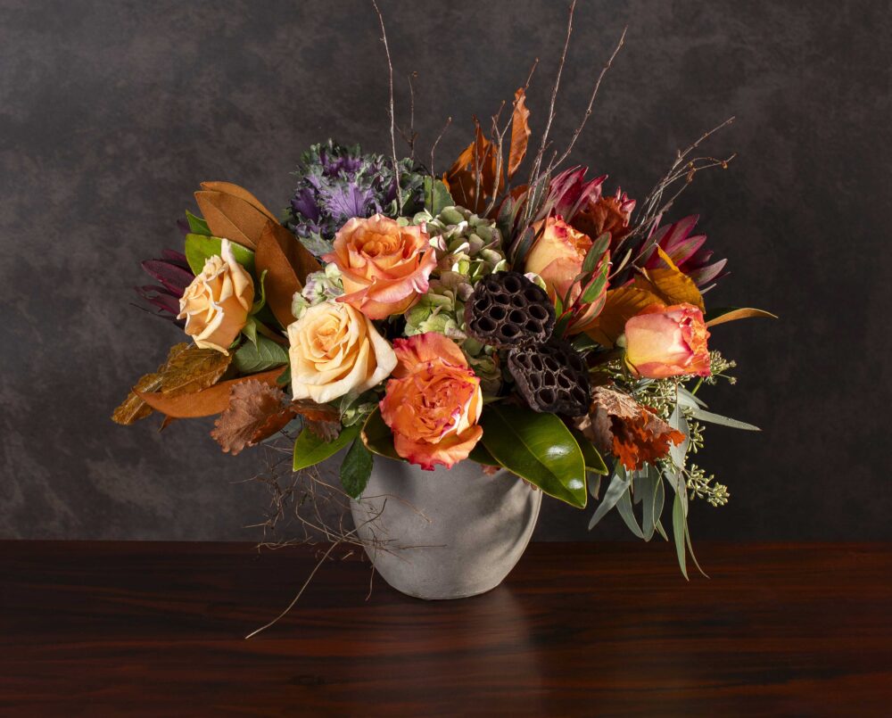 A fresh peach, coral, burgundy and purple mix of autumn seasonal flowers, brown leaves and texture in a concrete pot, delivered.