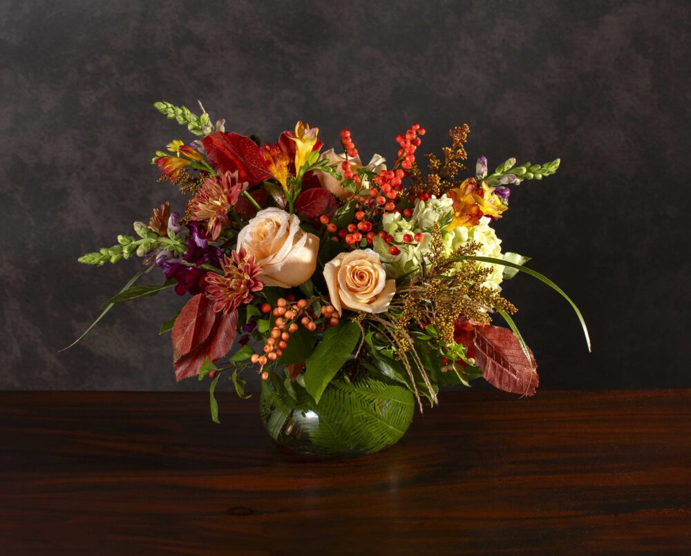 A vibrant crimson red, gold, peach and green mix of autumn seasonal flowers in a glass vase, delivered.