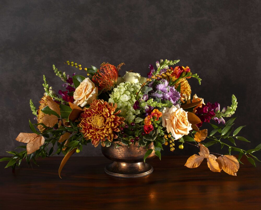 A deep orange, peach, purple, green, seasonal flower arrangement in a copper pedestal vase, delivered, thanksgiving