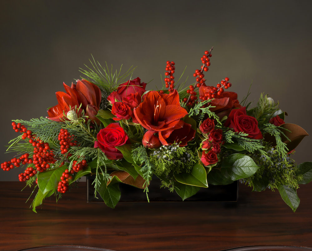 A bright red and green holiday centerpiece with red amaryllis, roses and ilex berry in lush evergreen and magnolia in a black ceramic rectangular vase, delivered, Christmas.