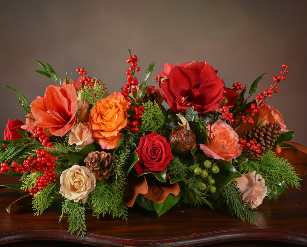 A bright, red, orange, peach, holiday, flower centerpiece with roses, amaryllis, ilex berry, hypericum, pinecones, magnolia, evergreens, delivered, holiday, Christmas.