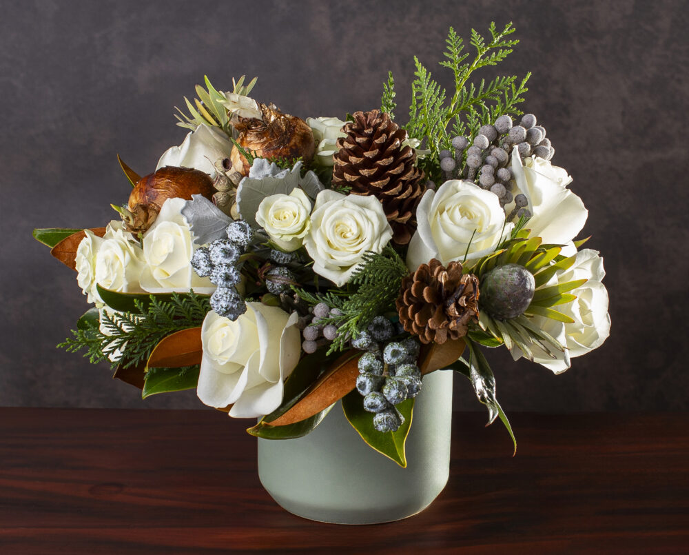 A seasonal, white, silver and green flower arrangement with roses, brunia, magnolia, pinecone, evergreen in a green vase, delivered, holiday, christmas