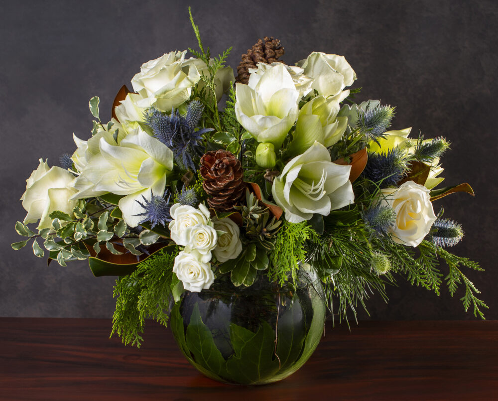 a refreshing white and blue flower arrangement with amaryllis, blue thistle eryngium, evergreens, magnolia in a glass vase with a leaf wrap for delivery, holiday.