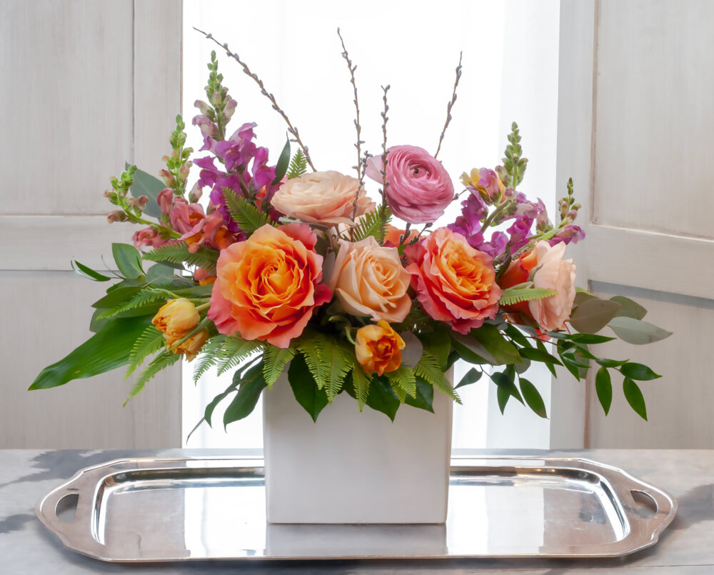 A bright and colorful winter arrangement, with seasonal flowers, roses, ranunculus, peach, pink and green, delivery