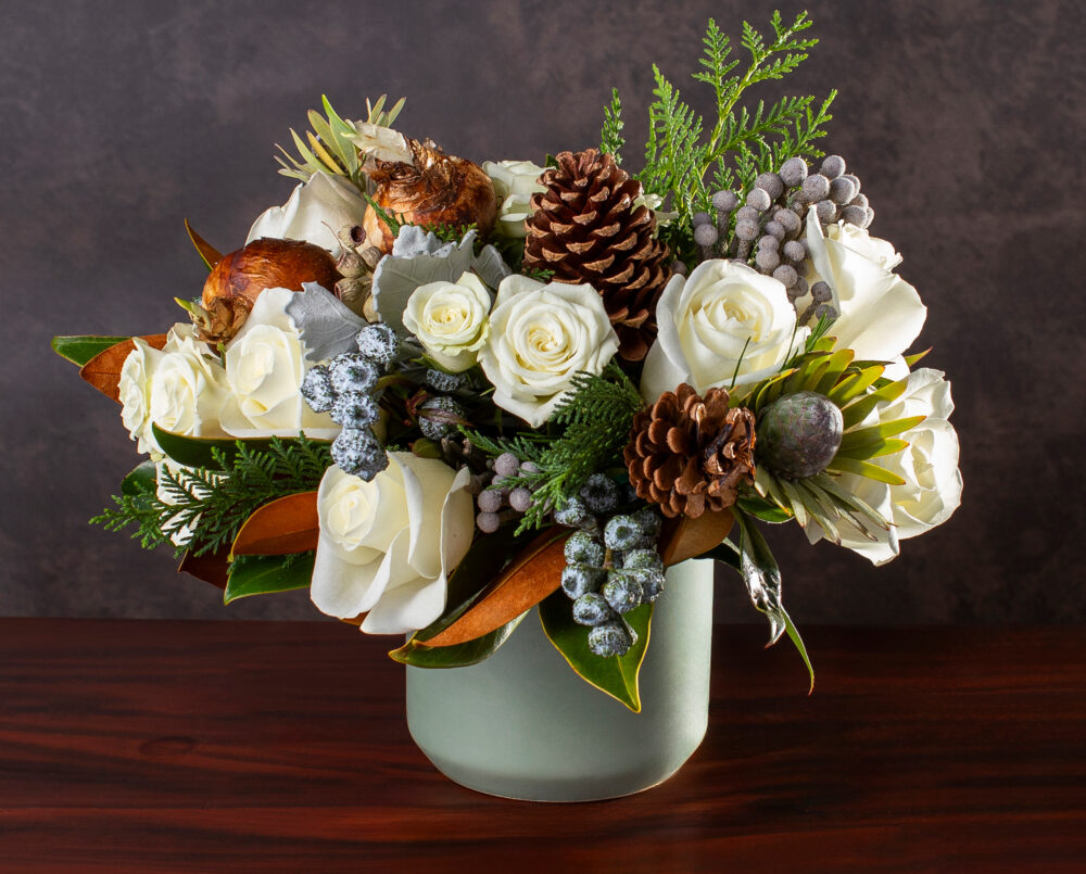 A fresh, white and green floral arrangement, with seasonal roses, brunia, pinecones, magnolia and evergreens in a green ceramic vase, delivered, winter.