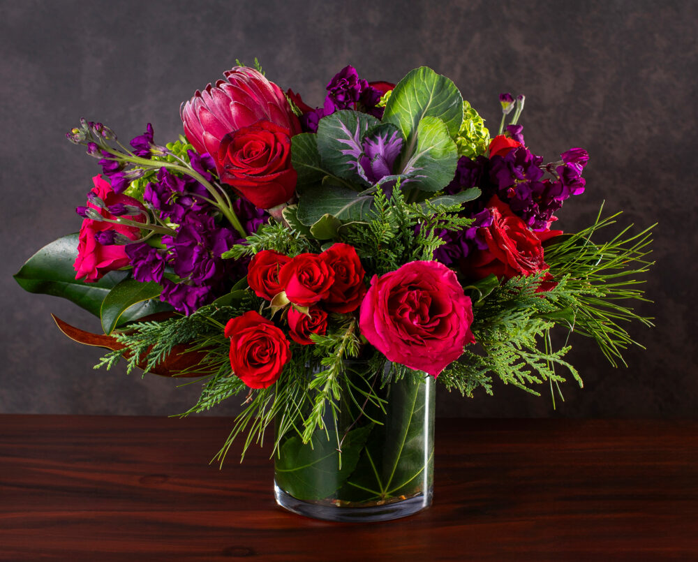A winter seasonal flower arrangement in bright colors of purple, pink and green in a clear glass vase with a leaf wrap, delivered.