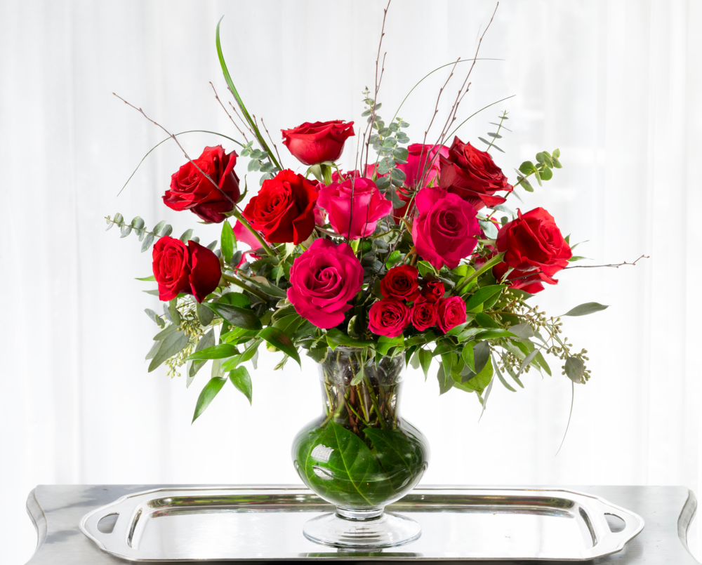 Dora is a tall and showy valentine flower arrangement in shades of red and pink in a glass vase with a leaf wrap.