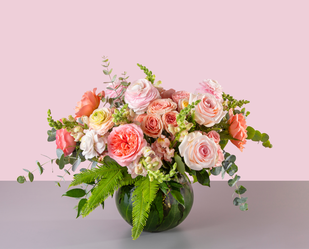 Elorie is a soft peach and pink arrangement with garden roses, ranunculus and snap dragons in a glass bowl vase.