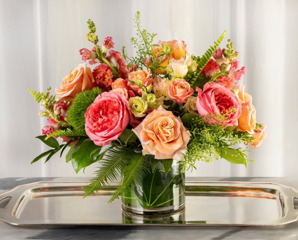 Marnie is a fresh coral and peach spring arrangement with unique roses, snap dragons and trachelium in a clear glass vase.
