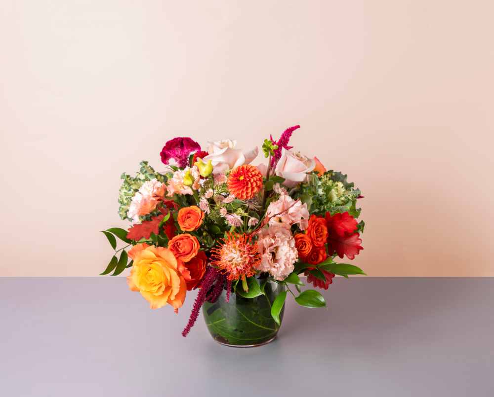 Henri is a bright orange and pink fall flower arrangement in a glass vase with a leaf wrap.