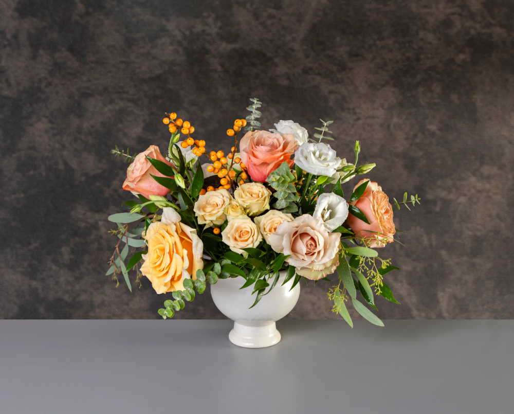 A fresh floral arrangement of peach, blush, cream and soft yellow roses with seasonal berries and greenery in a white pedestal compote vase.