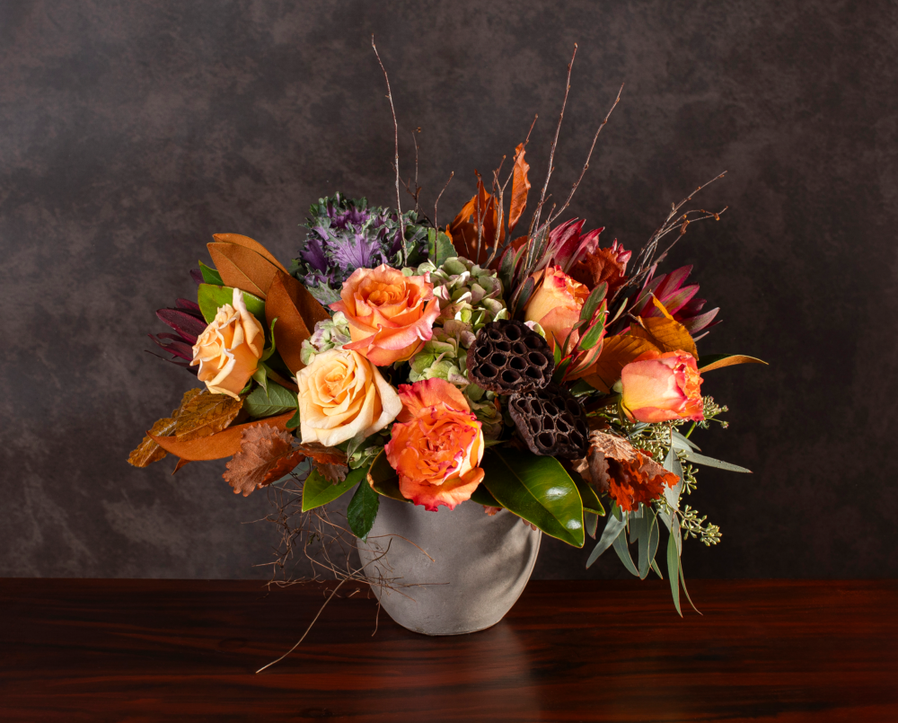A fresh fall floral arrangement of burnt orange, peach, burgundy and purple flowers with roses, autumn foliage, lotus pods and seasonal greenery in a textured stone compote vase.
